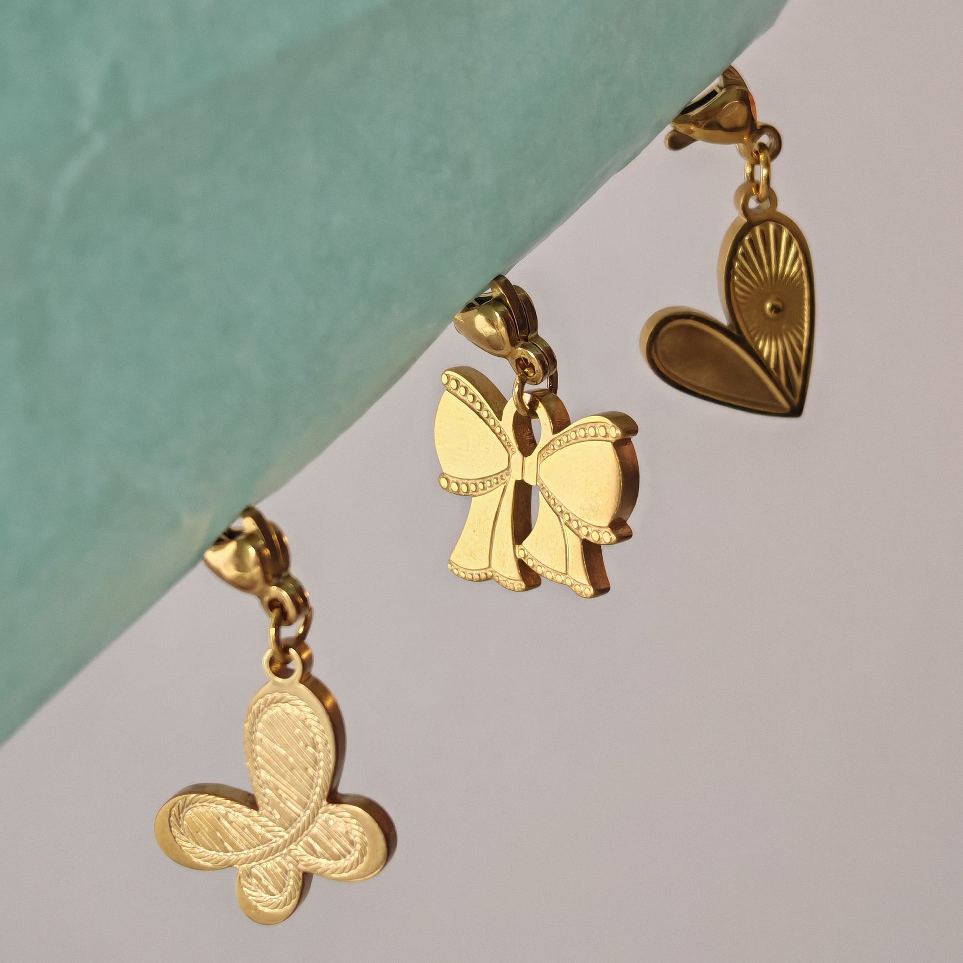 Trei charms aurii lucioase cu design delicat: fluture texturat, fundiță și inimă gravată.